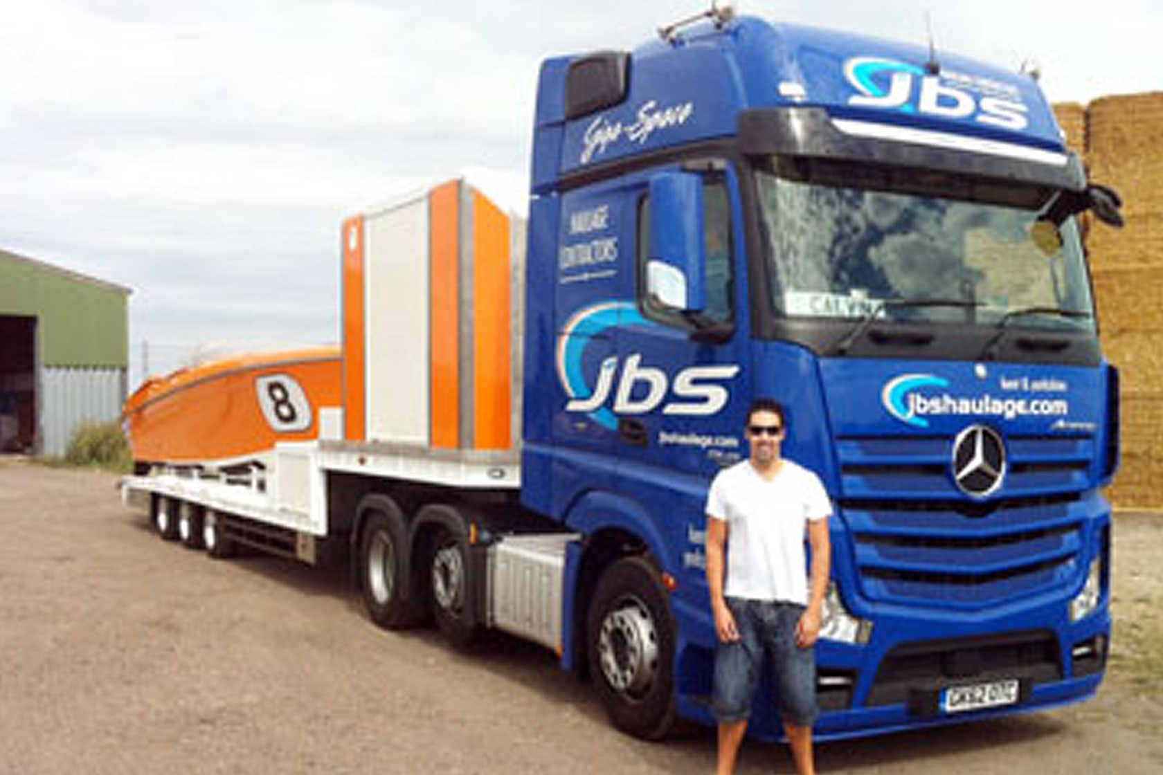 a photo of Calvin stood next to a. blue JBS truck that has a flatbed on it carrying a speedboat 